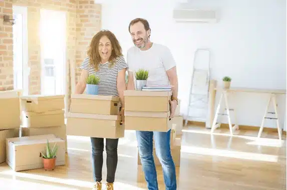A couple smiling while carrying moving boxes in an empty room.