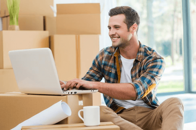 Young man searching on a laptop surrounded by moving boxes.