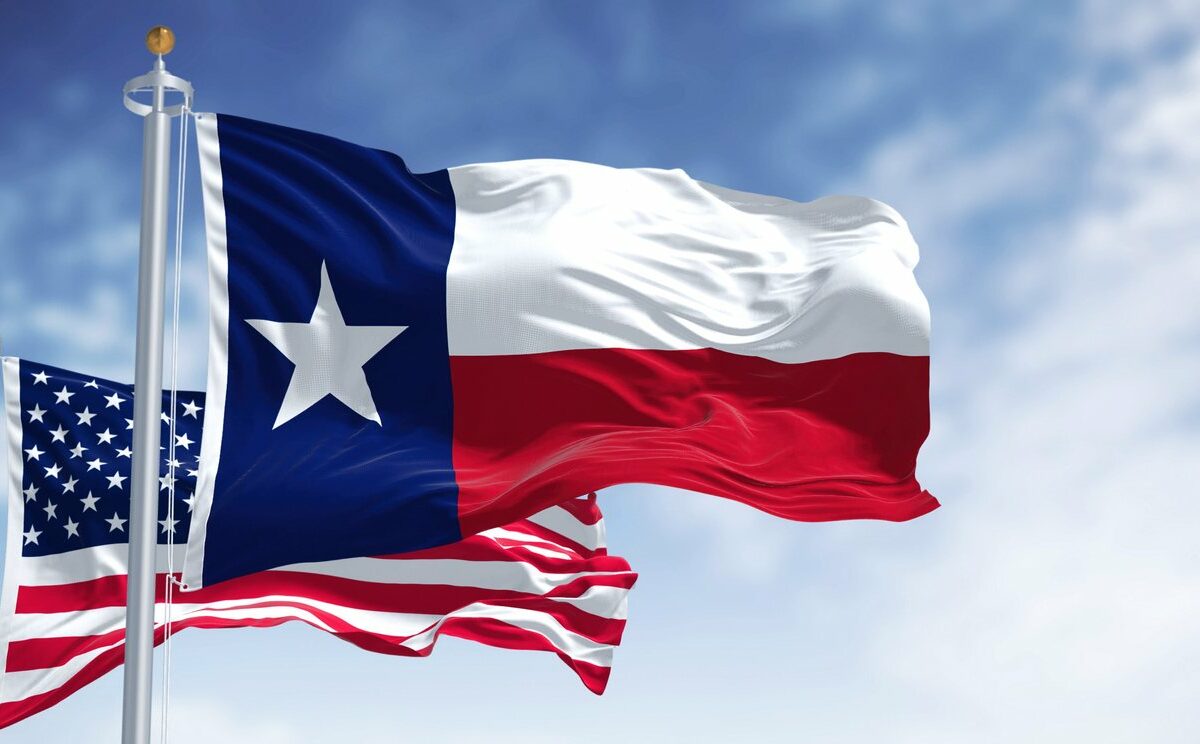 The Texas flag flies in front of the United States flag against a blue sky.