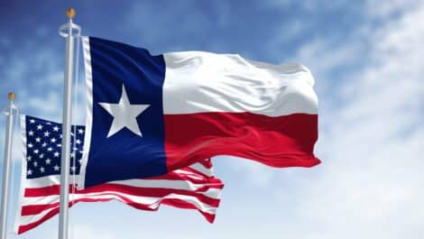 The Texas flag flies in front of the United States flag against a blue sky.
