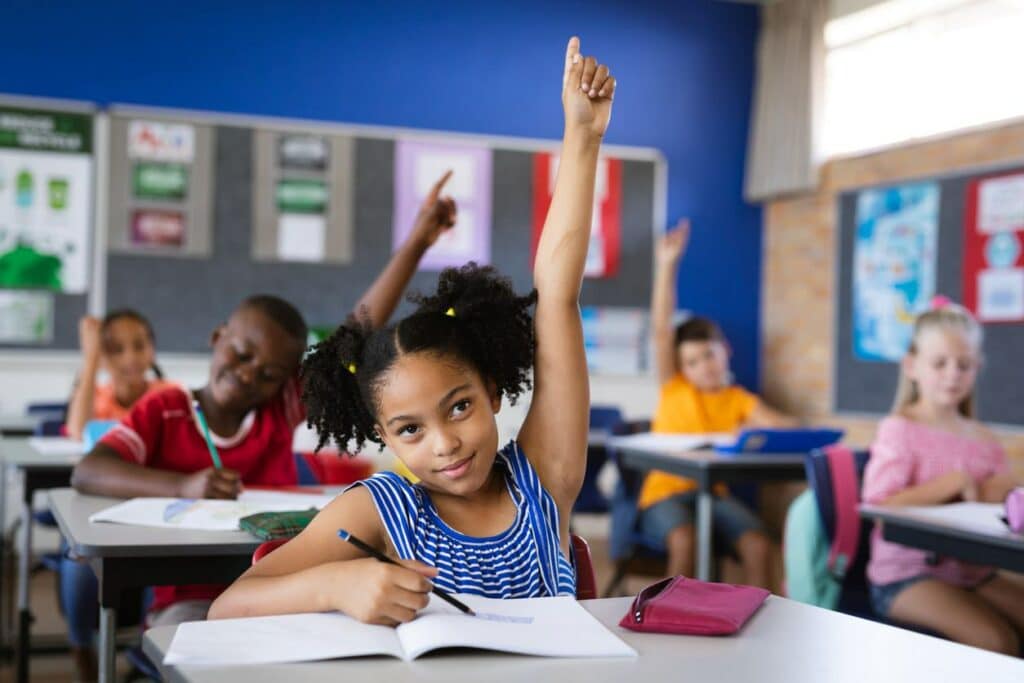 A young girl sitting at a school desk is raising her hand to answer a question.