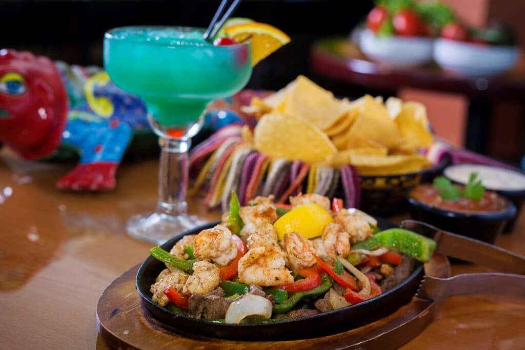 a plate of fajitas with a blue margarita and corn chips in the background.