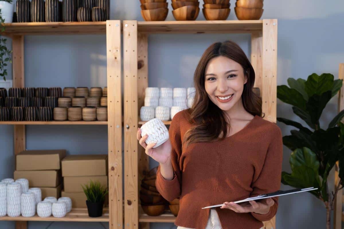 Ceramist smiles while holding a clay pot and a clipboard with a shelf full of pottery behind her