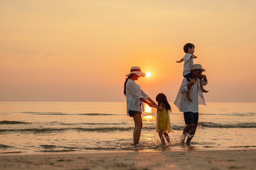 Mother holds daughter’s hands and father holds son on shoulders while playing on the beach