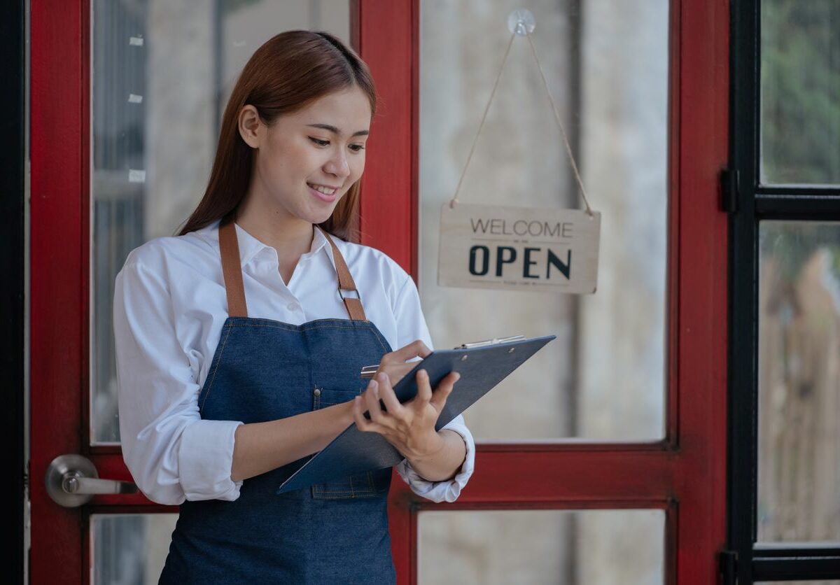 a woman in an apron smiles as she holds a clipboard and writes on a tablet