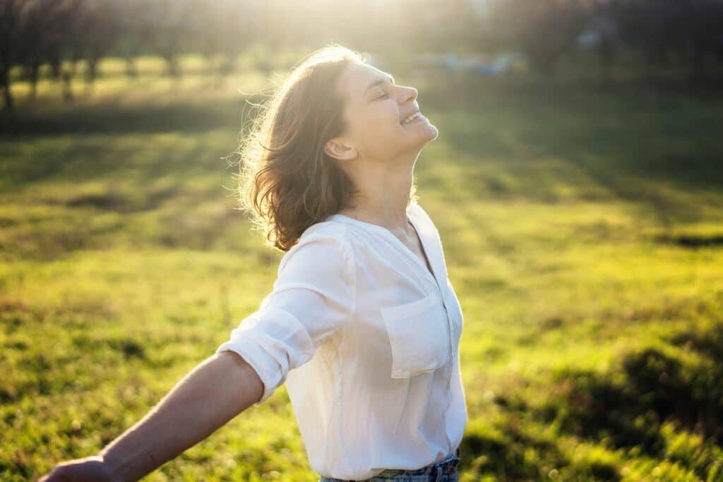 A woman in a white shirt smiles and basks in the sun while standing in a green field at sunset